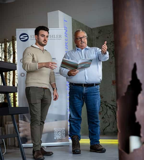 Zwei Fachkräfte stehen in einem modernen Innenraum und besprechen ein Projekt; ein älterer Mann zeigt mit der Hand nach vorne, während er eine Broschüre hält, und ein jüngerer Kollege folgt aufmerksam seinen Anweisungen, im Hintergrund ist ein Steinkühler-Display zu sehen – Steinkühler Wasser & Wärme GmbH & Co. KG
